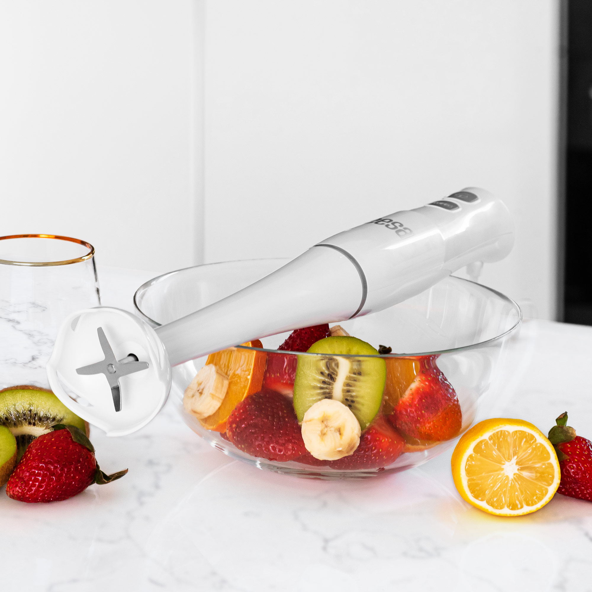 Biały blender ręczny leży nad szklaną miską wypełnioną pokrojonymi owocami, takimi jak truskawki, kiwi i banany.