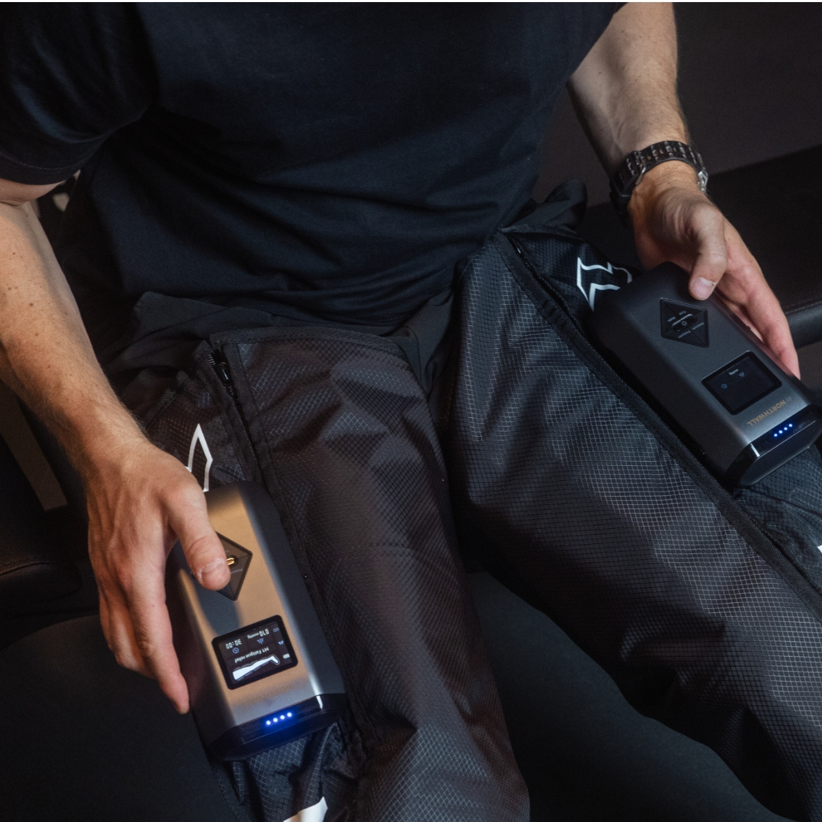 Un homme en vêtements noirs est assis et tient deux contrôleurs gris pour bottes de compression.