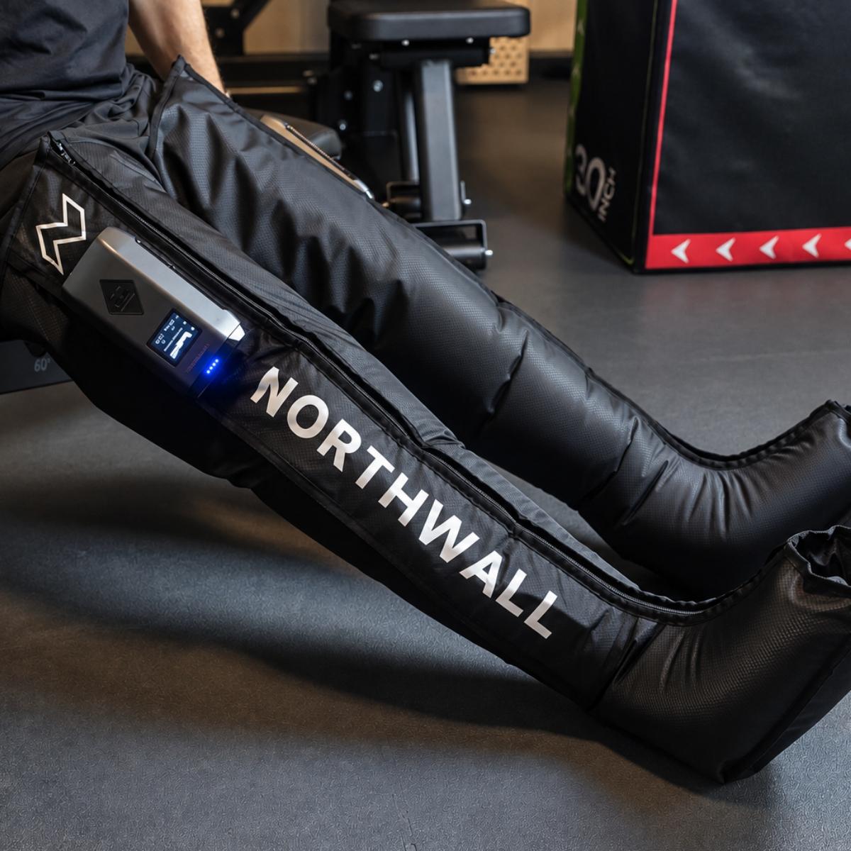 Une personne porte des bottes de compression noires Northwall avec un écran dans une salle de sport.
