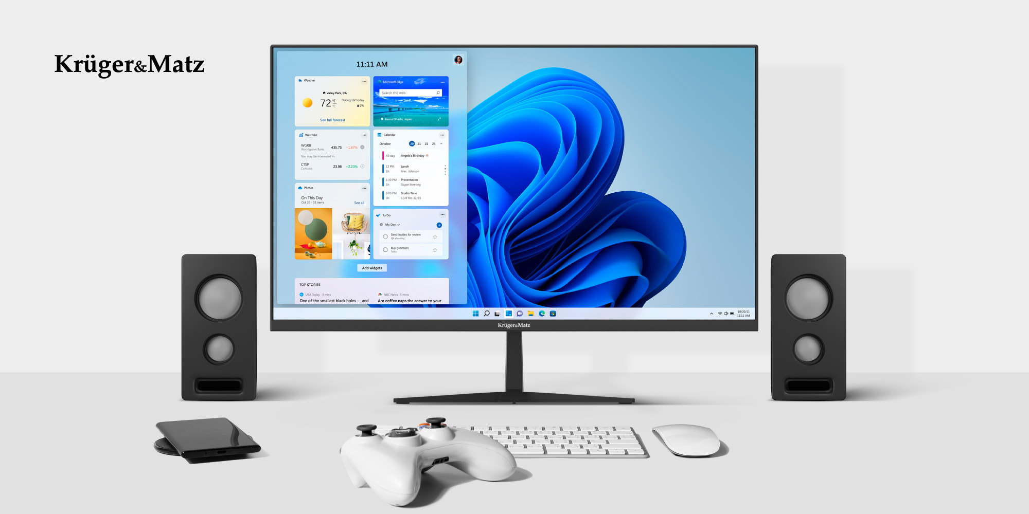 Monitor z pulpitem Windows 11, w otoczeniu dwóch głośników, klawiatury, myszy, pada do gry i smartfona.