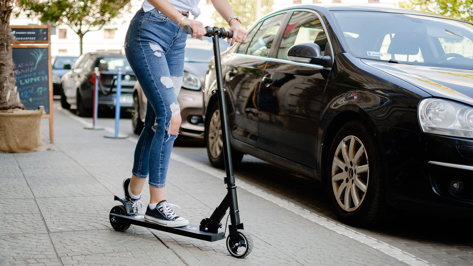 Kobieta w postrzępionych dżinsach jedzie na czarnej hulajnodze elektrycznej po chodniku obok zaparkowanych samochodów.