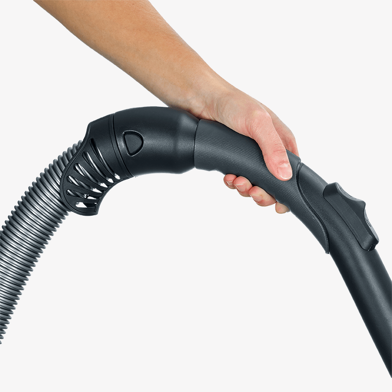 Una mano sostiene el mango negro y ergonómico de una aspiradora con manguera gris sobre fondo blanco.