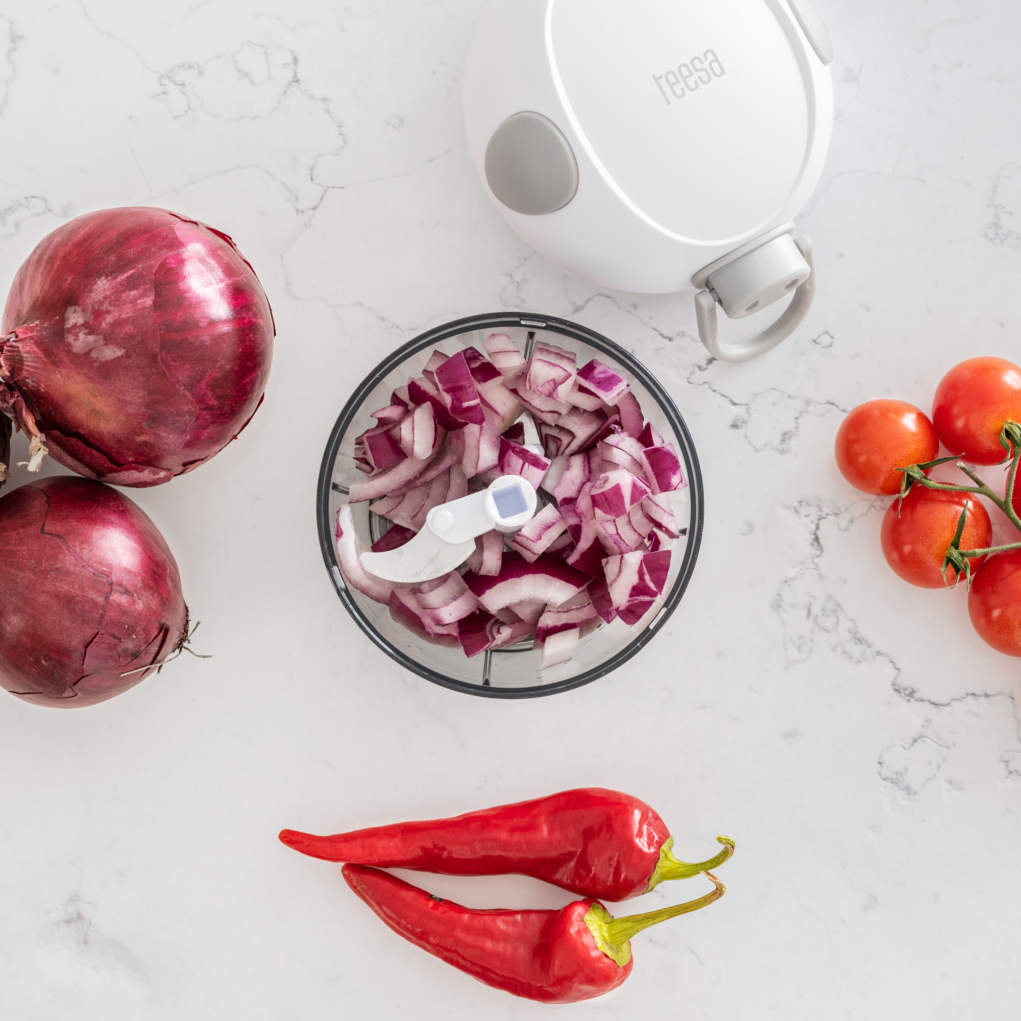 Biała siekaczka ręczna z posiekaną czerwoną cebulą, otoczona całymi cebulami, papryczkami chili i pomidorkami koktajlowymi.