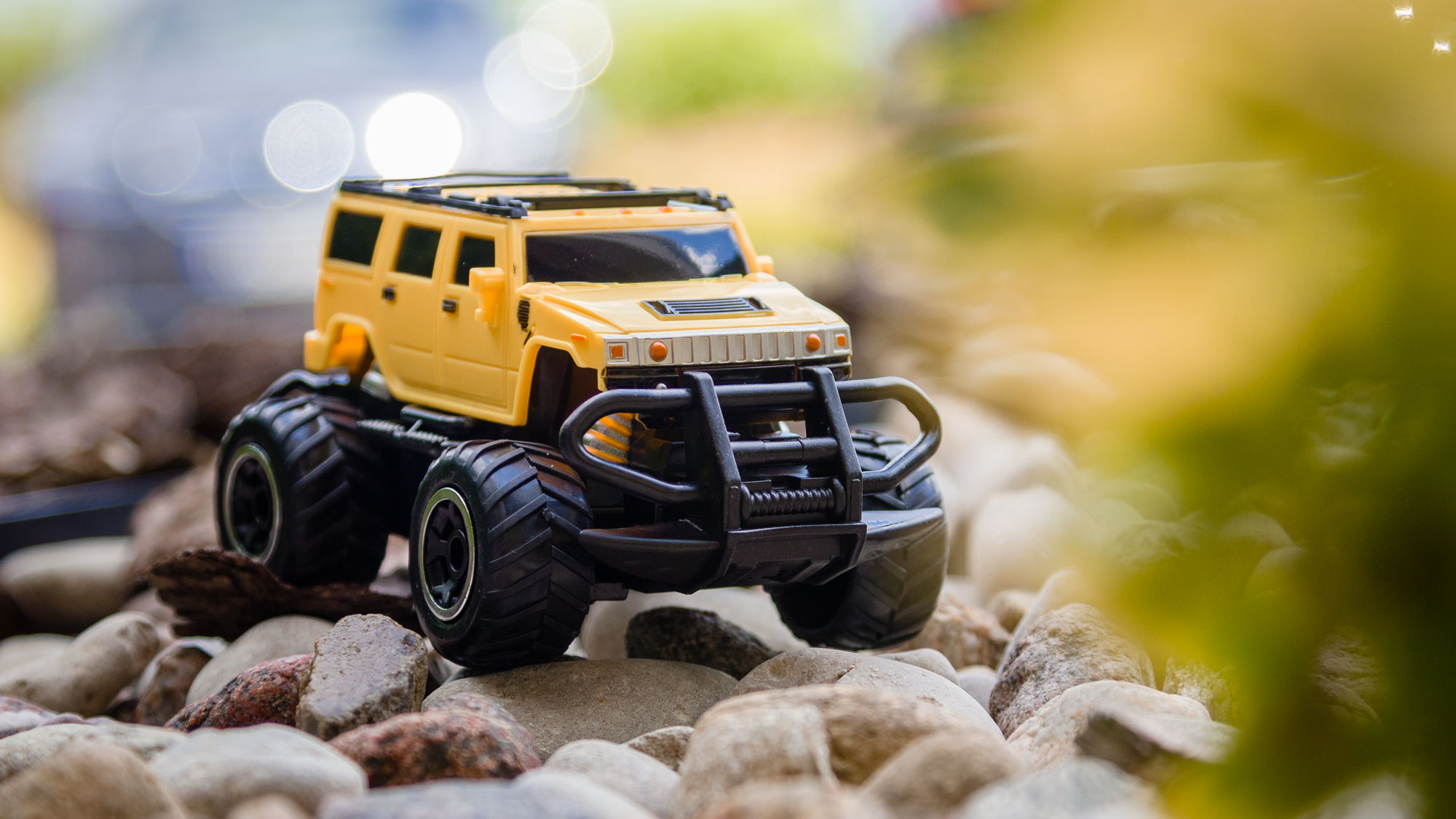 Żółty zabawkowy monster truck z dużymi czarnymi oponami jedzie po małych kamieniach na zewnątrz.
