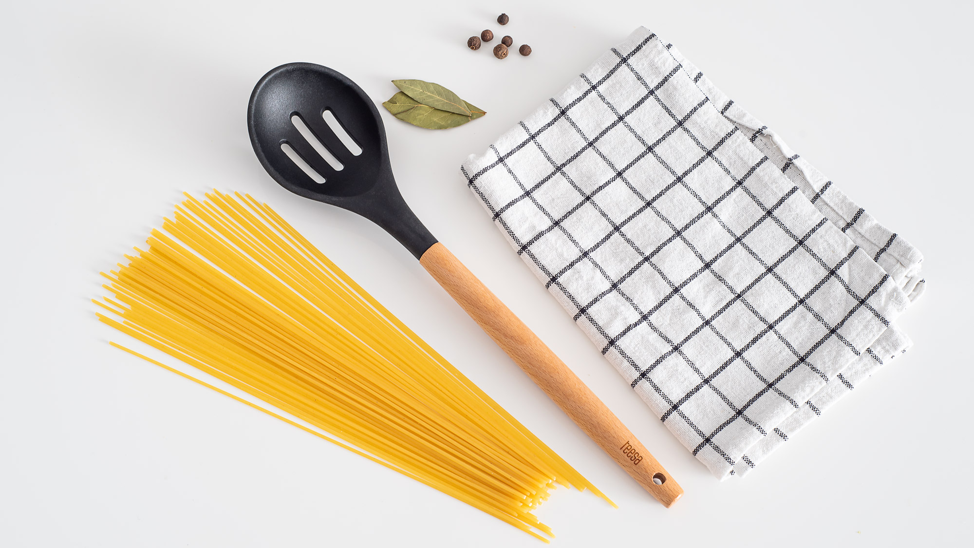 Spaghetti, czarna lyzka kuchenna, liscie laurowe, ziarna pieprzu i w kratke sciereczka na bialym stole.