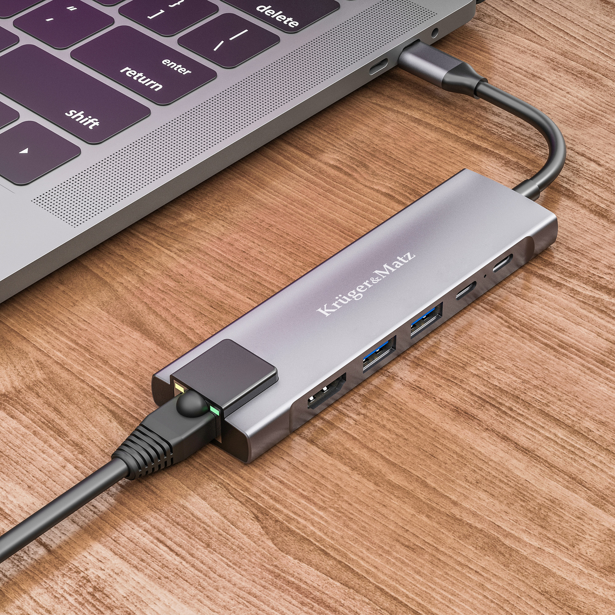 Szary hub USB-C jest podłączony do laptopa i kabla Ethernet na drewnianym stole.