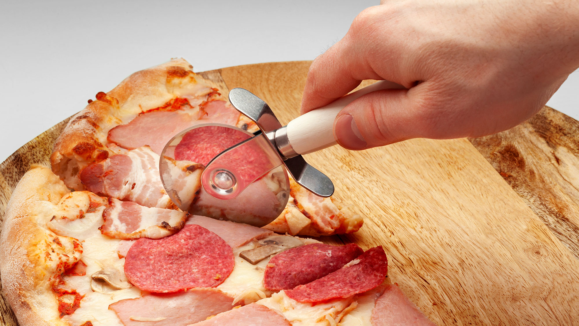 Ręka kroi pizzę z salami, szynką i pieczarkami na drewnianej desce za pomocą noża krążkowego.
