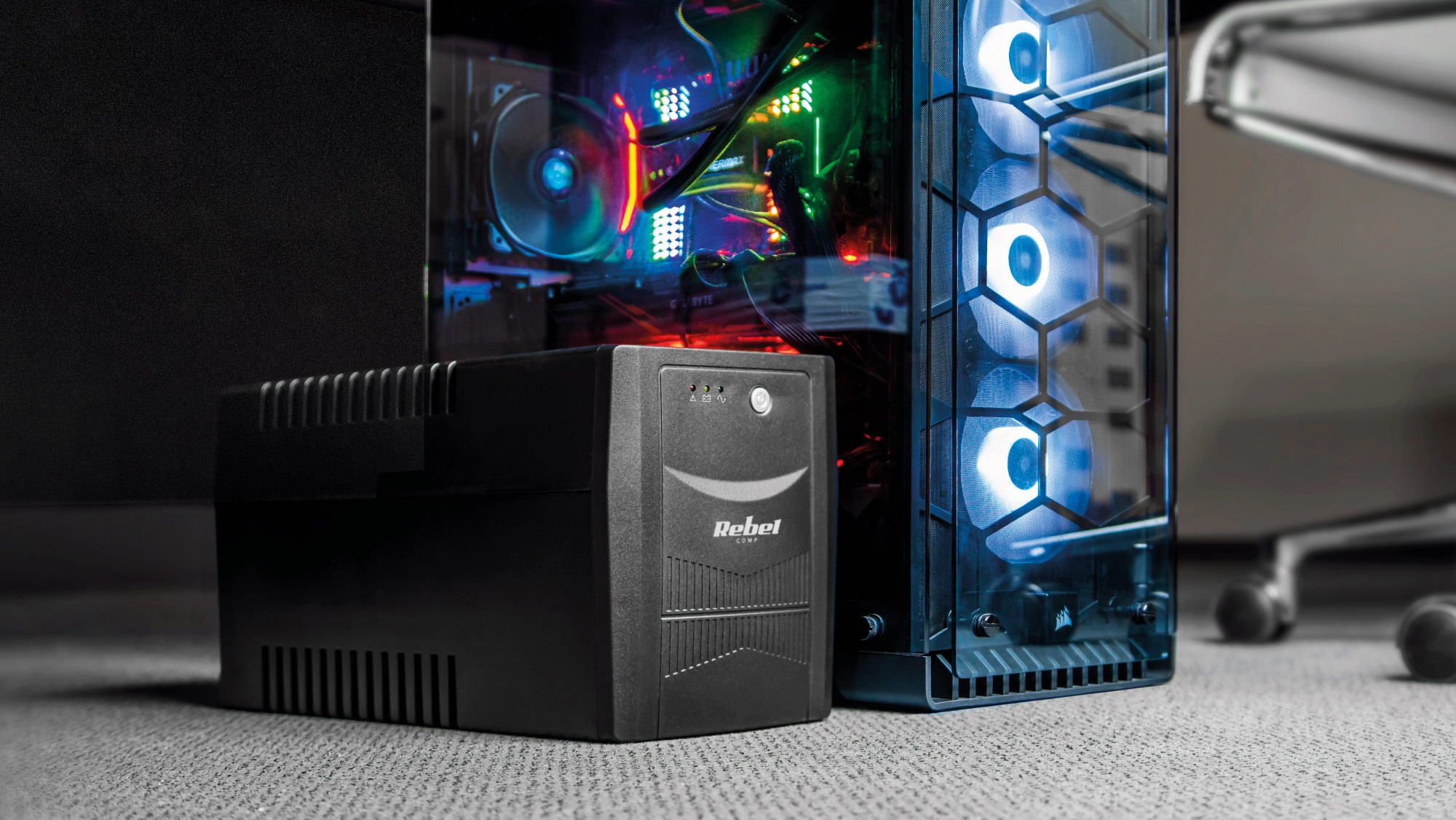 Czarny zasilacz awaryjny UPS Rebel stoi na dywanie przed obudową PC z wentylatorami RGB.