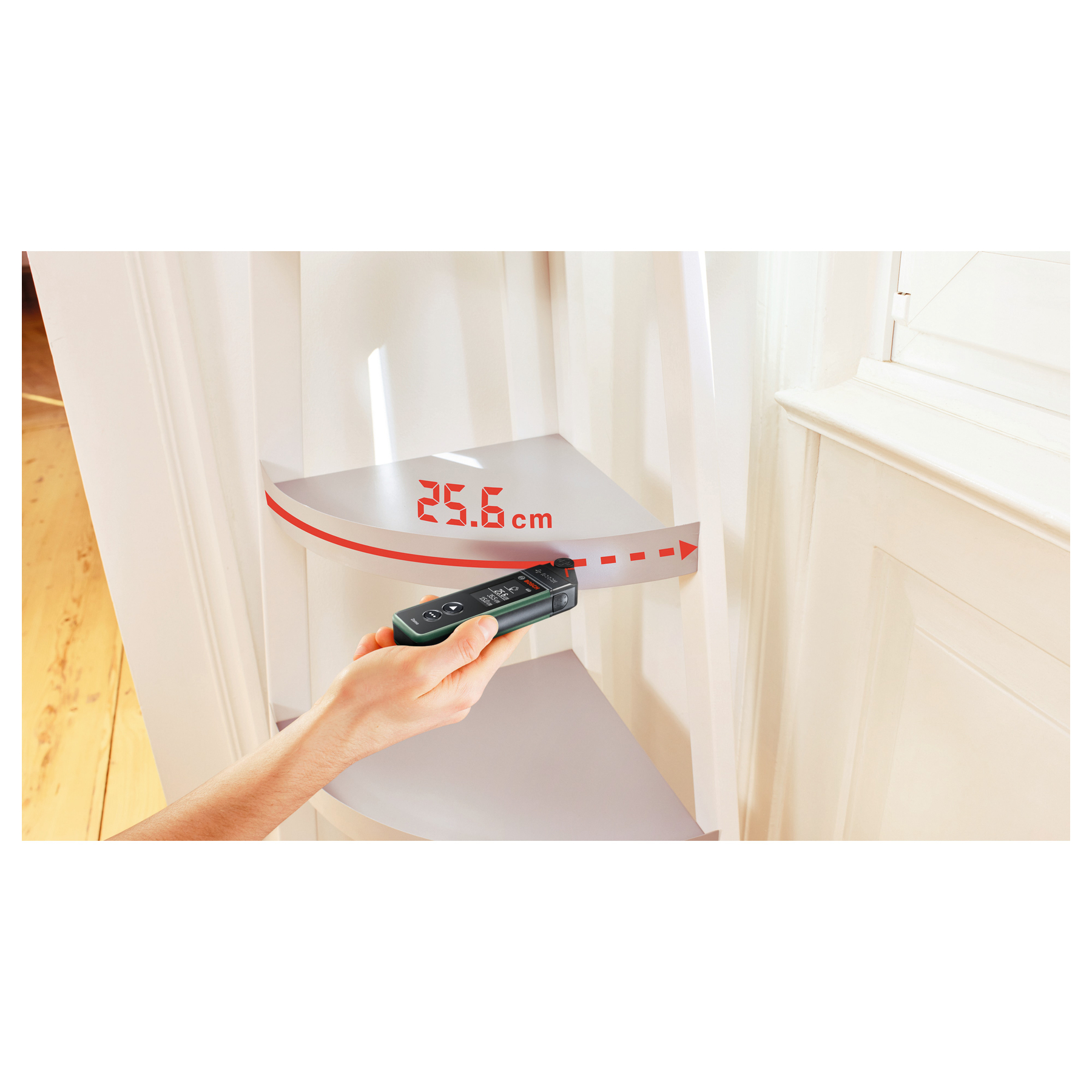 Hand hält ein Lasermessgerät vor ein weißes Eckregal, Display zeigt 25,6 cm.