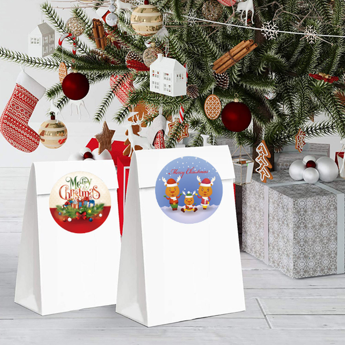 Zwei weiße Geschenktüten mit Weihnachtsaufklebern stehen vor einem geschmückten Baum und Geschenken.