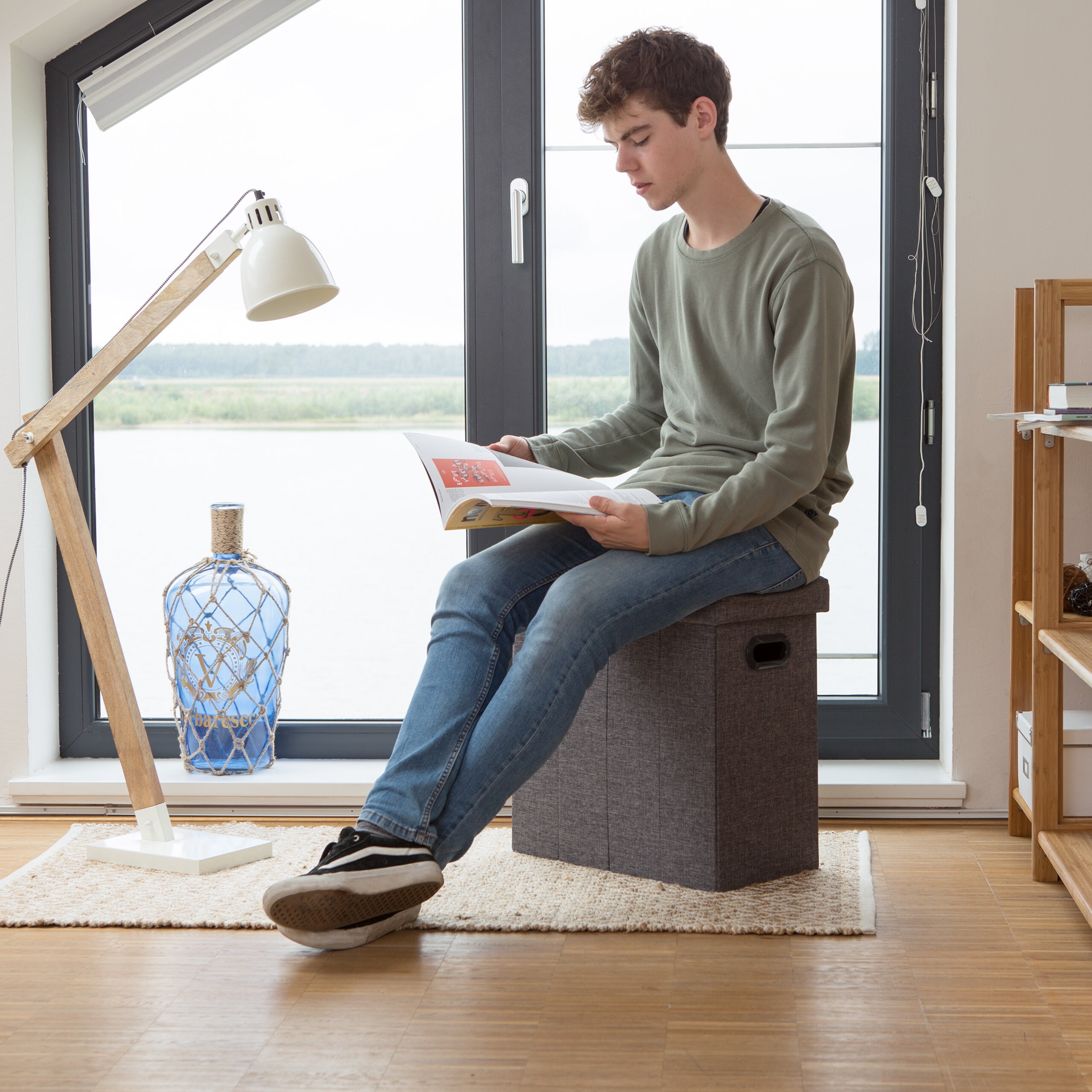 Ein junger Mann sitzt auf einem Hocker vor einem großen Fenster und liest ein Magazin.