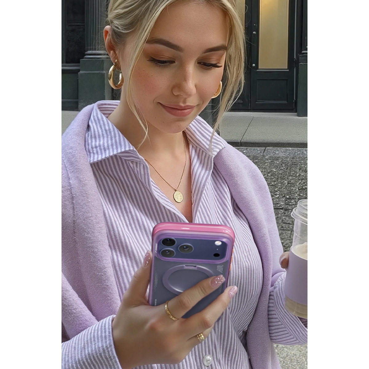 Mor ve beyaz çizgili gömlekli sarışın bir kadın, bir binanın önünde akıllı telefon ve kahve tutuyor.