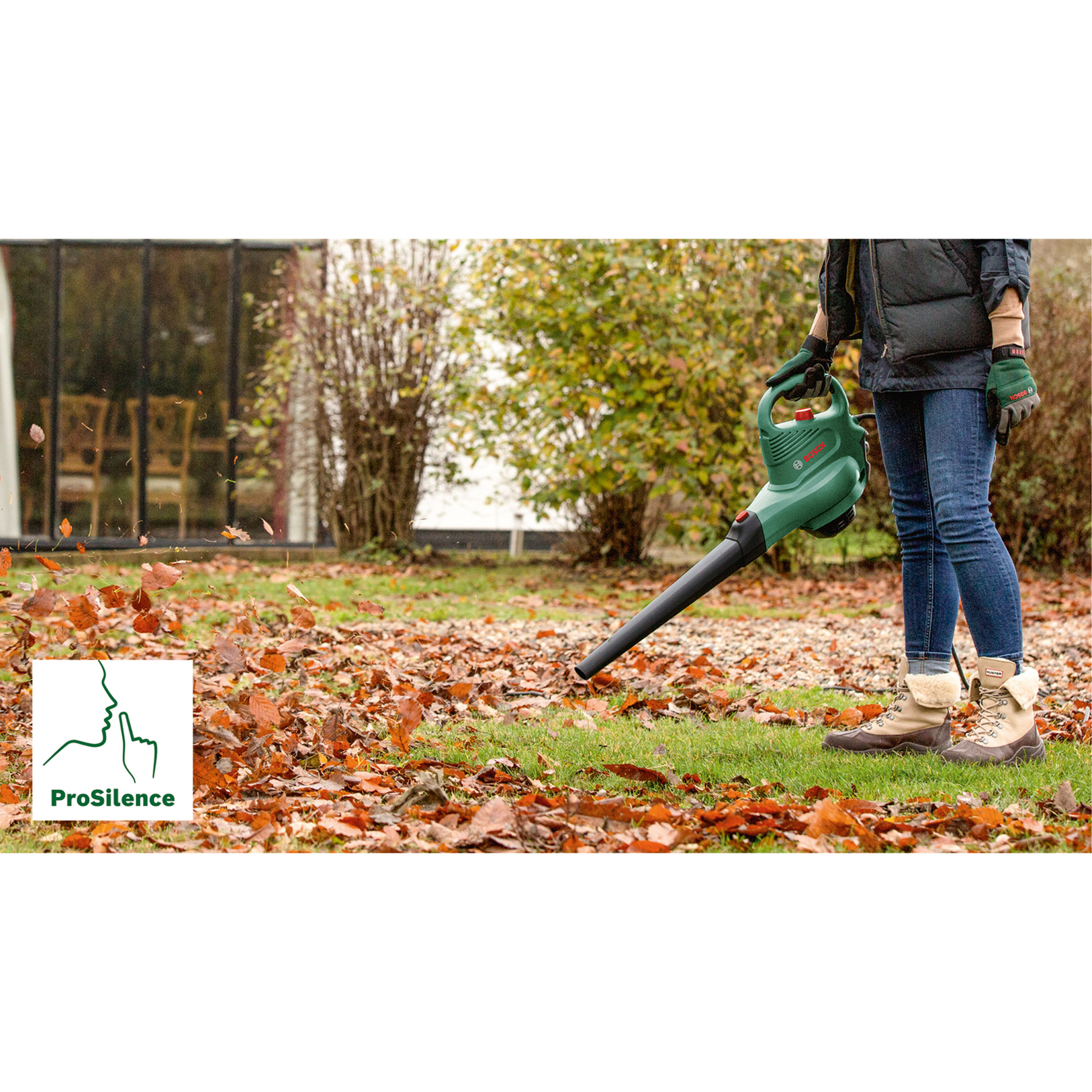 Eine Person benutzt einen grünen Laubbläser, um herbstliches Laub von einem Rasen zu entfernen.