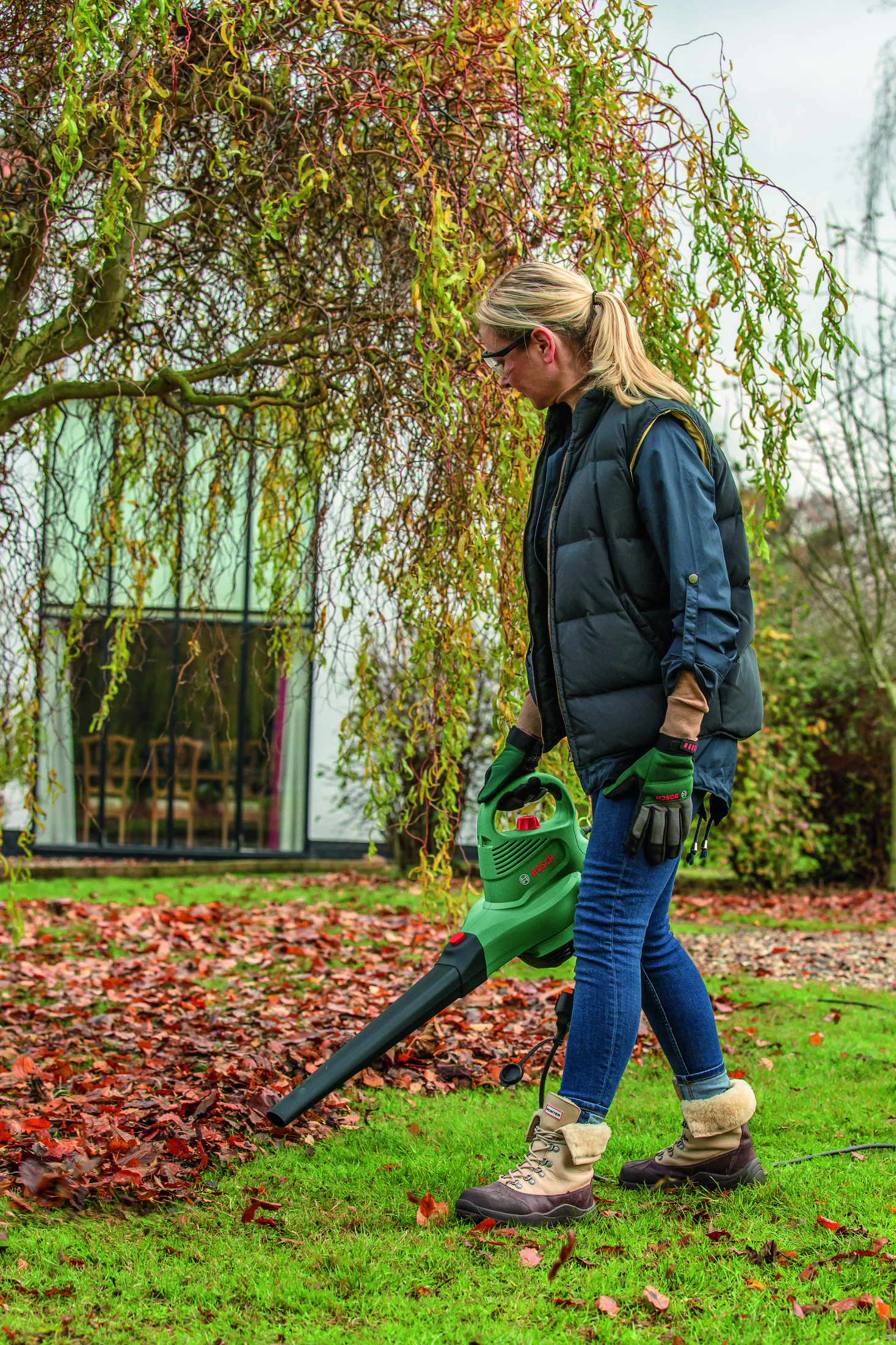 Eine Frau in einer Weste hält einen grünen Laubsauger im Garten mit Herbstlaub.