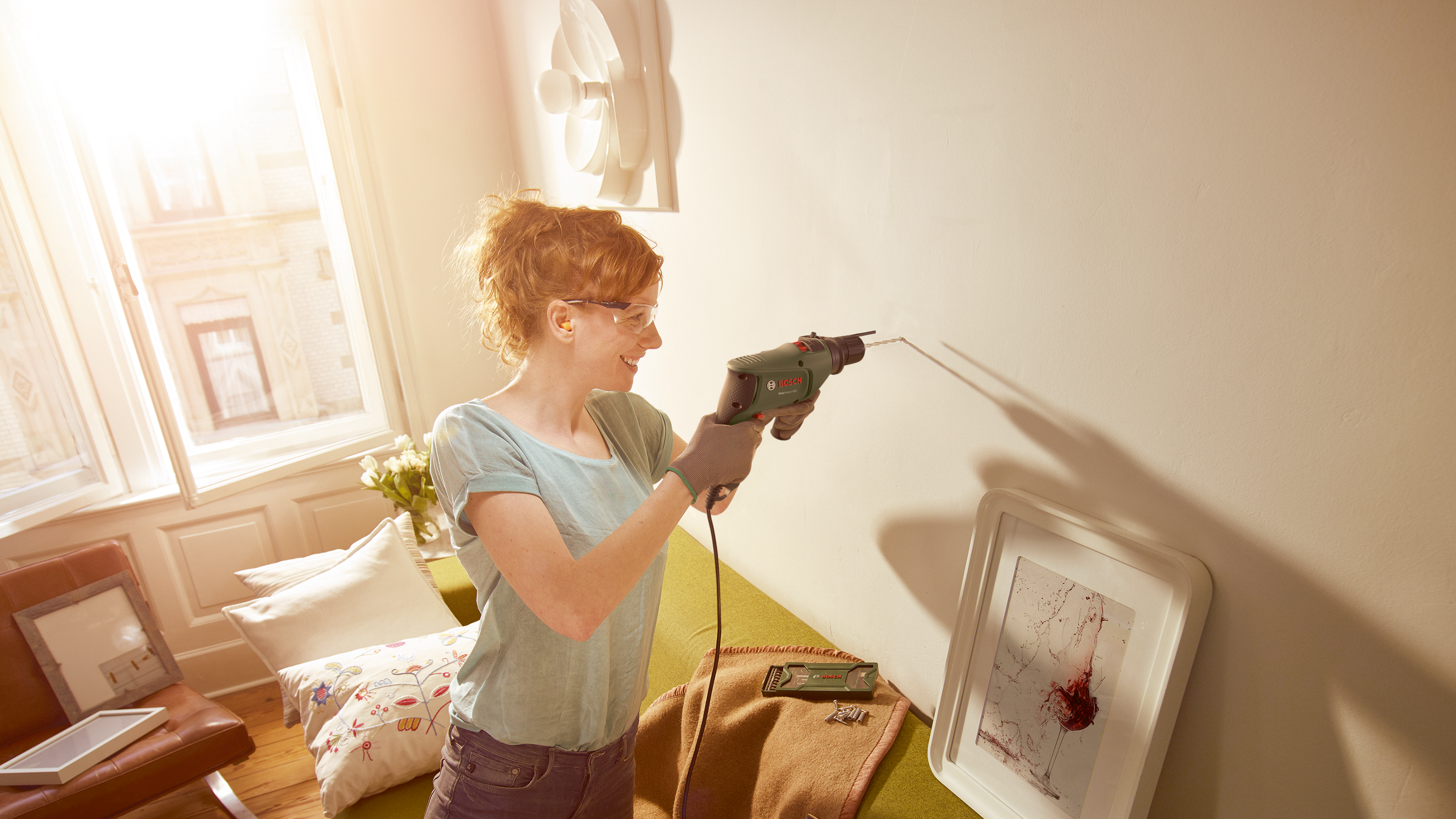 Eine Frau bohrt in einer sonnigen Wohnung ein Loch in die weiße Wand.