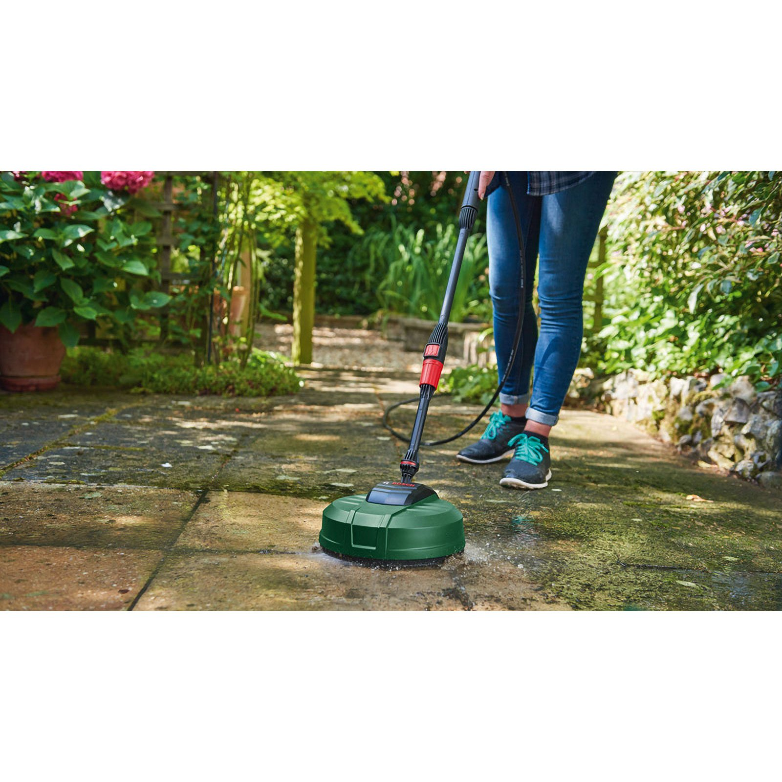 Eine Person reinigt Steinplatten im Garten mit einem grünen Terrassenreiniger vor üppigen Pflanzen.