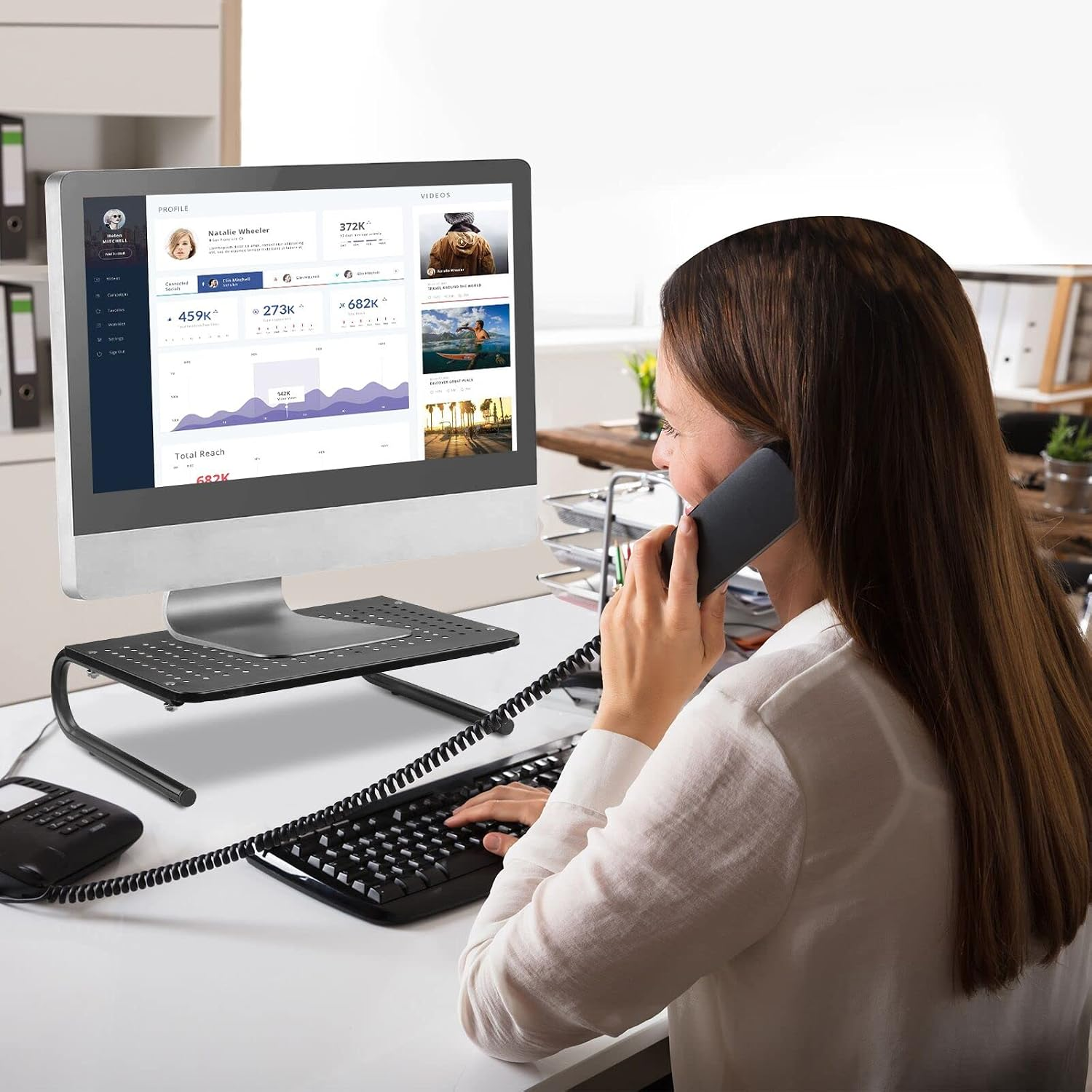 Eine Frau in weißer Bluse telefoniert am Schreibtisch vor einem Computer mit Statistiken.