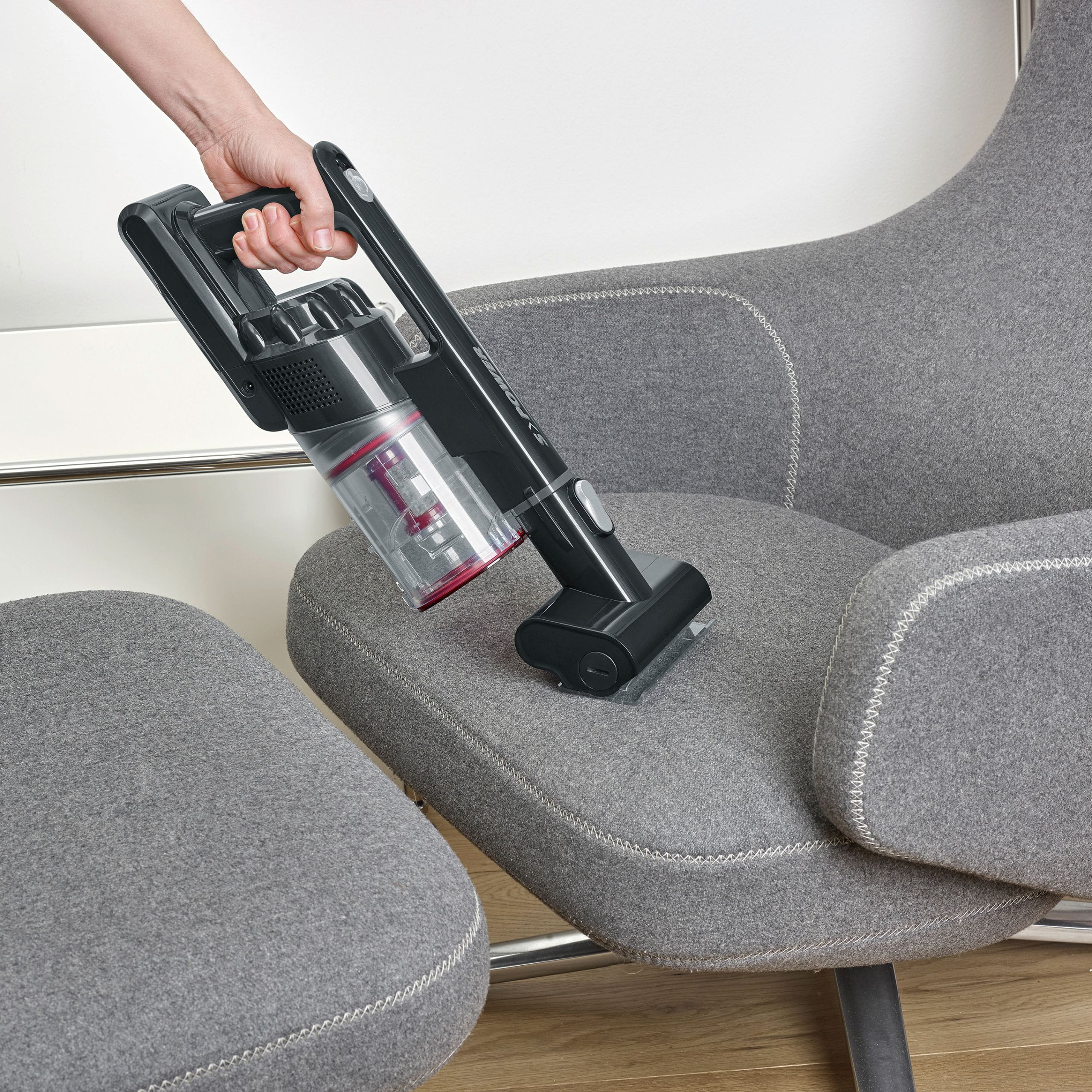 Eine Hand hält einen grauen Handstaubsauger, der einen grauen Sessel reinigt.