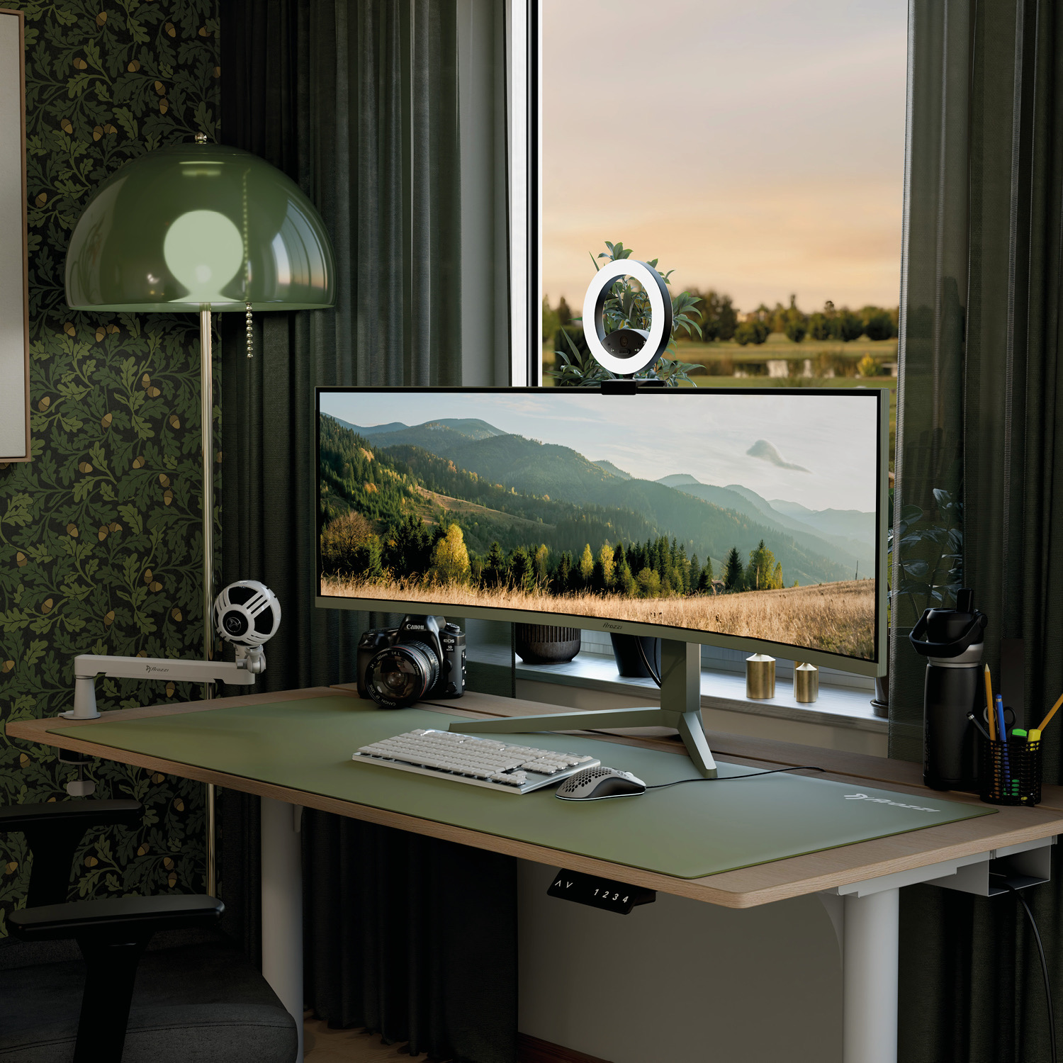 Ein grüner Schreibtisch mit Monitor, Kamera, Ringlicht und Lampe vor einem Fenster mit Landschaftsblick.