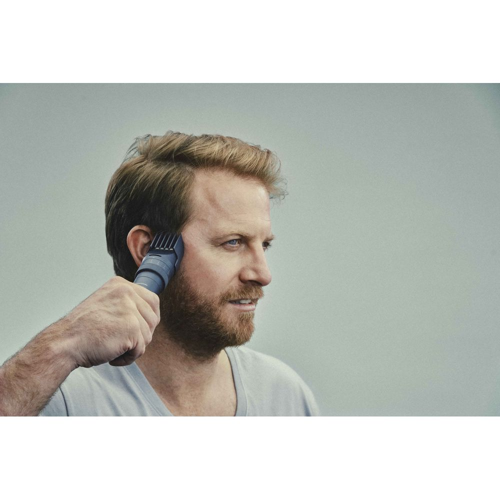 Ein Mann mit Bart trimmt sein Haar mit einem grauen Haarschneider vor grauem Hintergrund.