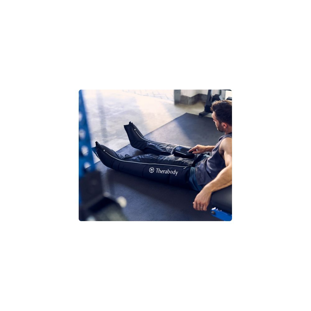 Un uomo siede in palestra e aziona stivali a compressione Therabody sulle sue gambe.