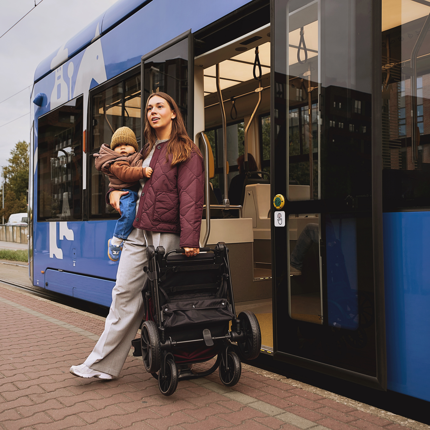 Una mujer con un bebé y un carrito plegado sube a un tranvía azul y blanco en una estación.