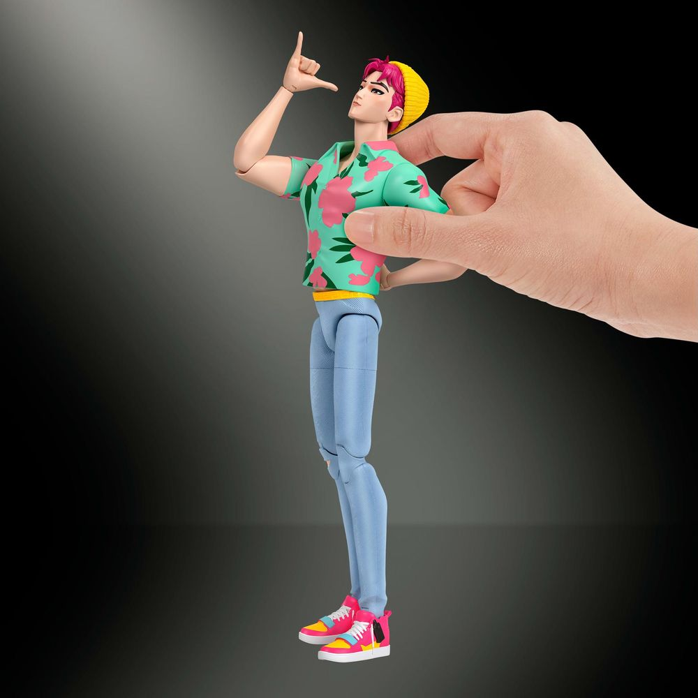 Una mano sostiene una muñeca de moda masculina con pelo rosa, ropa colorida y gorro amarillo ante fondo oscuro.