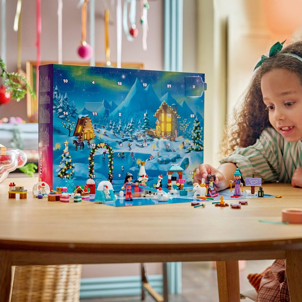 Dziewczynka przy stole bawi się figurkami Lego Friends przed dużym kalendarzem adwentowym z zimowym krajobrazem.