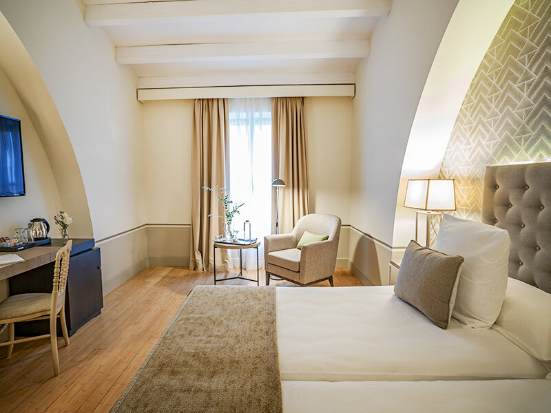Acogedora habitación de hotel con una cama en primer plano, sillón junto a la ventana y suelo de madera.