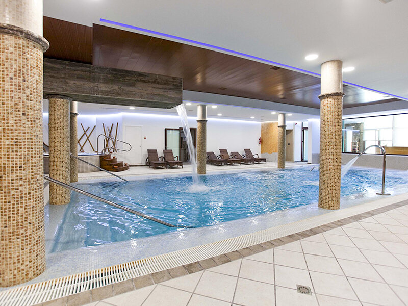 Piscina de spa interior con agua azul, cascada, pilares de mosaico y tumbonas al fondo.