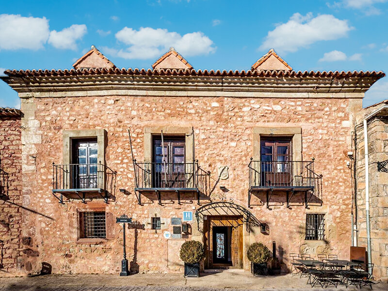 Un edificio de piedra rústica con tres balcones, puertas de madera y un cielo azul al fondo.