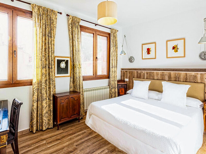 Un dormitorio luminoso con suelo de madera, una cama grande con cojines blancos y ventanas con cortinas estampadas.