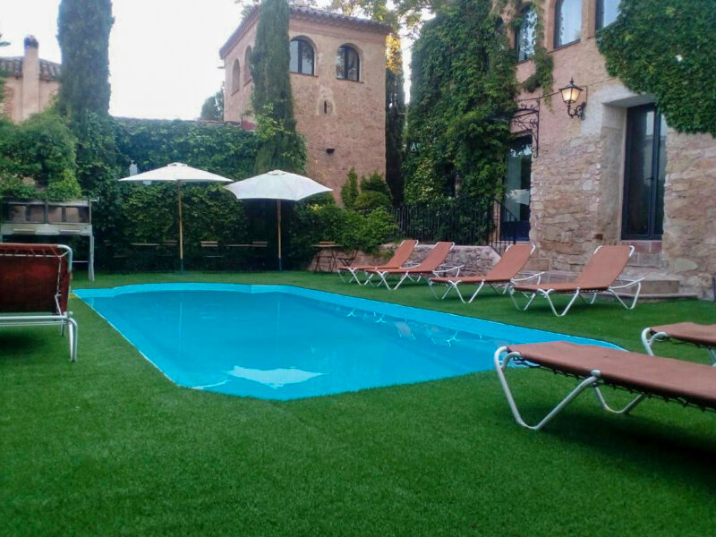 Una piscina azul en césped verde, rodeada de tumbonas y edificios históricos de piedra.
