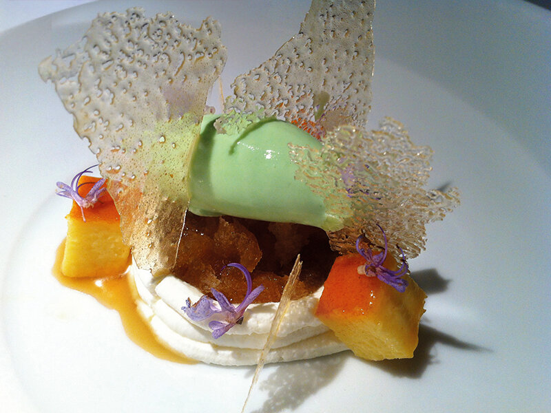 Postre artístico con helado verde, merengue, fruta caramelizada y flores comestibles en un plato blanco.