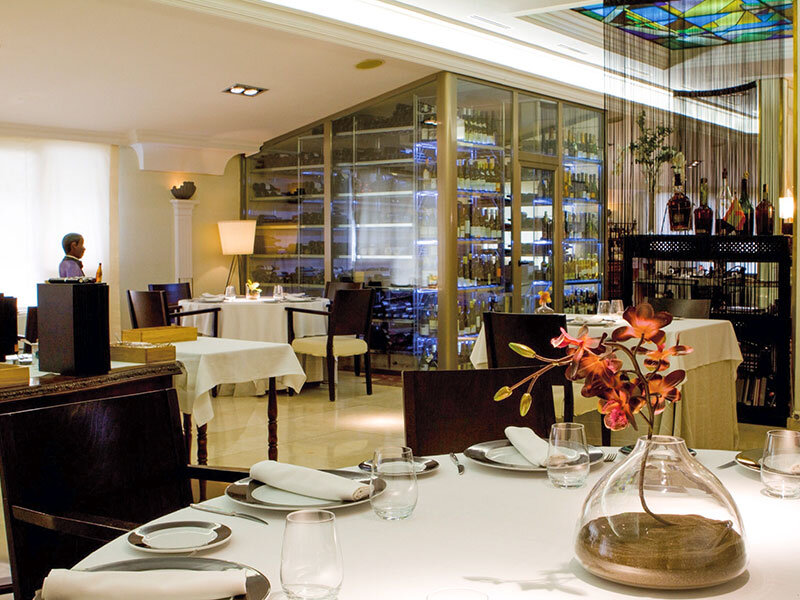 Un restaurante elegante con mesas blancas, sillas oscuras y un gran expositor de vinos de cristal al fondo.