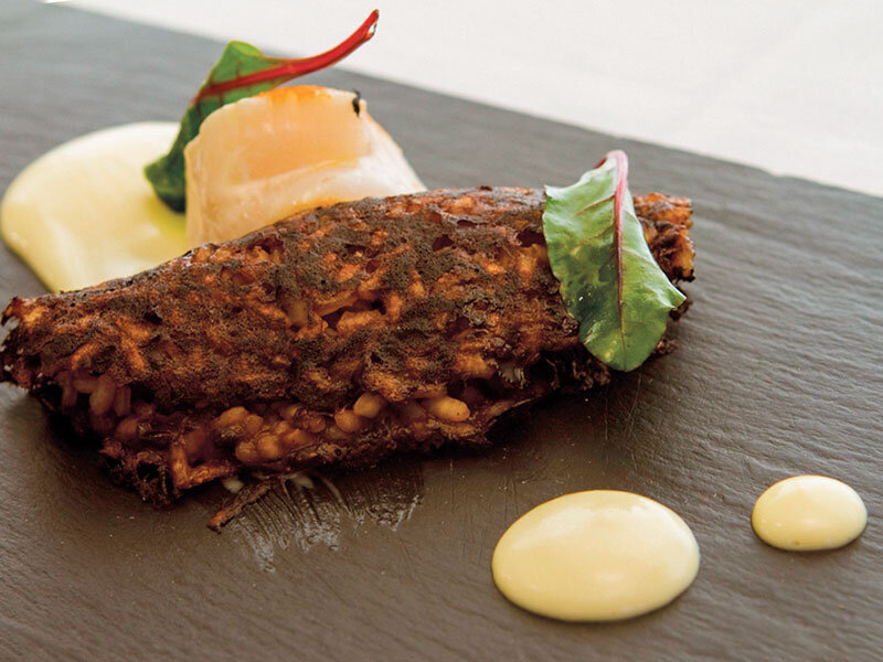 Plato crujiente en pizarra con vieira, hojas verdes y gotas de salsa amarilla.