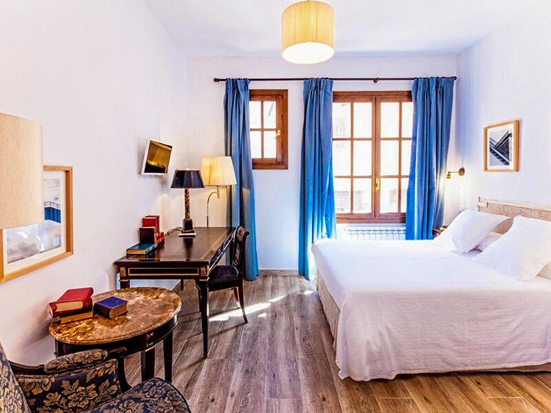 Un dormitorio luminoso con una cama doble, cortinas azules, un escritorio y suelo de madera.