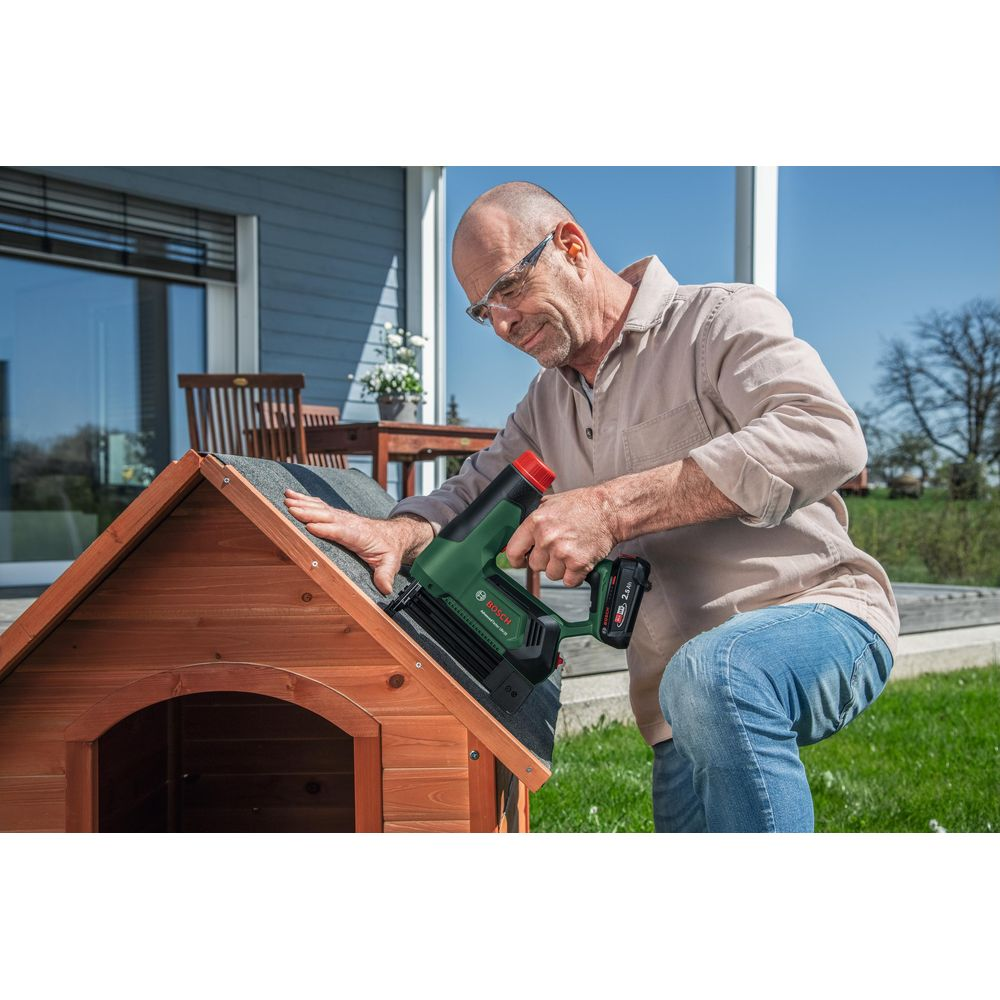 Ein Mann nutzt einen grünen Nagler am Dach einer Holzhütte im Garten.