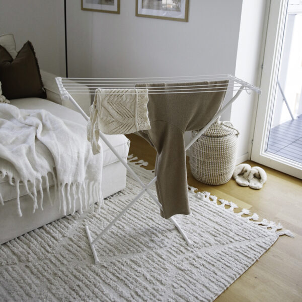 Un séchoir à linge blanc est posé sur un tapis à motifs dans une pièce lumineuse, avec des vêtements neutres.