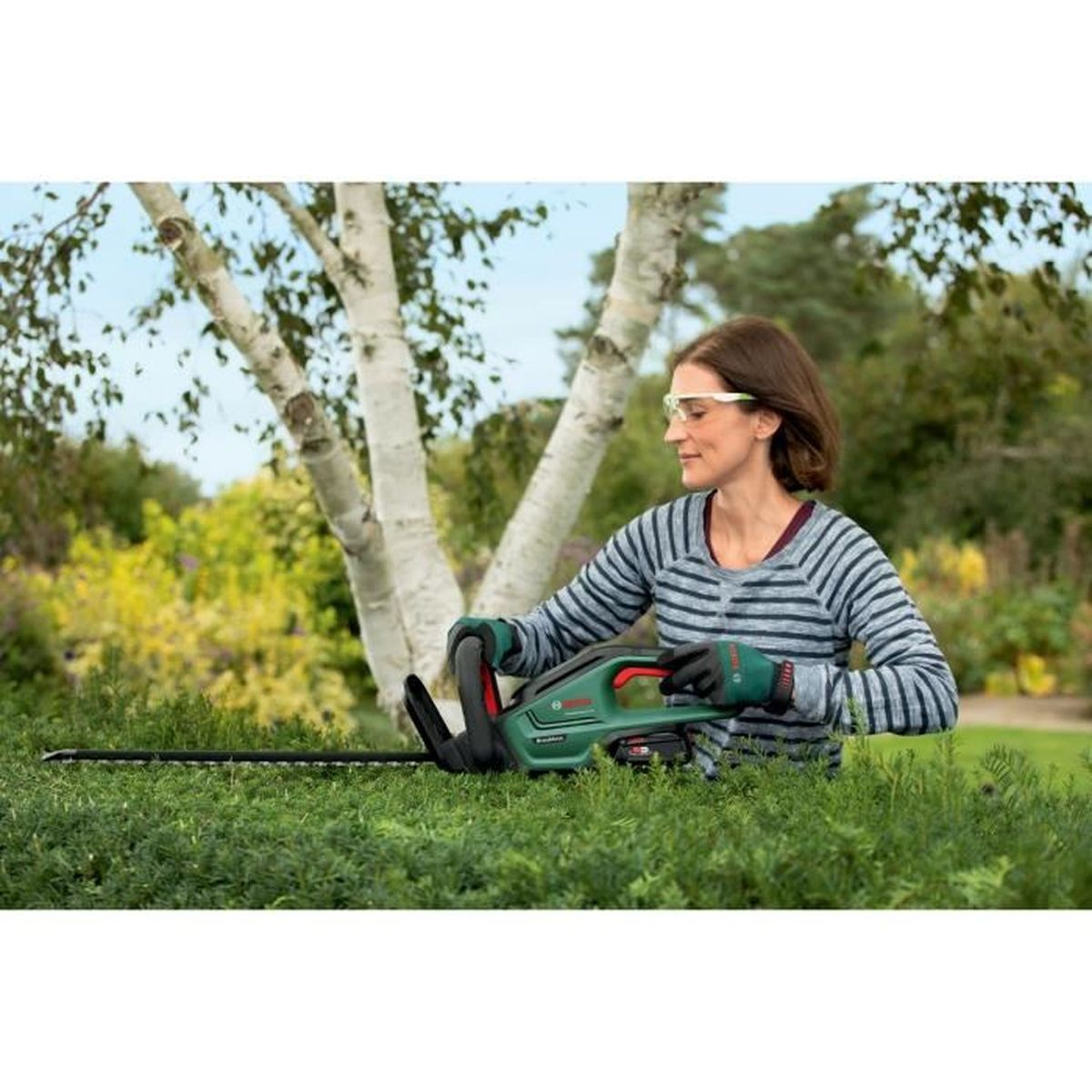 Eine Frau mit Schutzbrille und Handschuhen schneidet mit einer Akku-Heckenschere eine Hecke im Garten.
