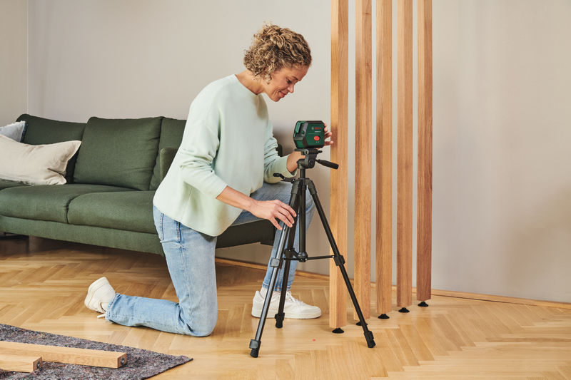 Eine Frau in Jeans kniet im Wohnzimmer auf dem Parkettboden und stellt einen grünen Laser auf einem Stativ ein.