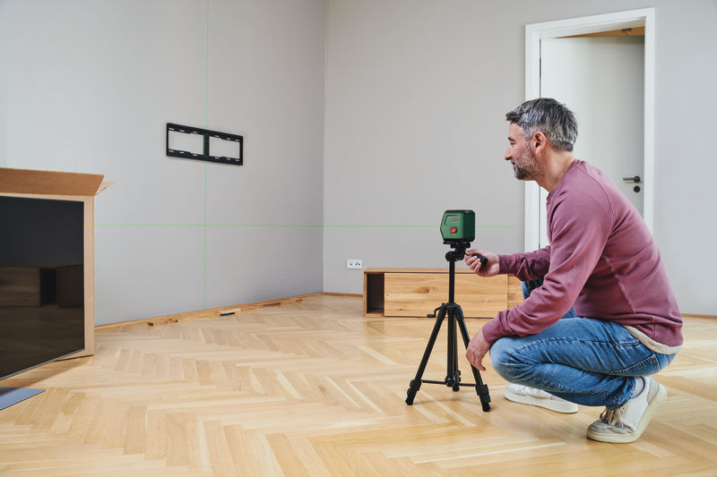 Ein Mann im rosa Pullover kniet und stellt einen grünen Laser-Level auf einem Stativ auf Holzfußboden ein.