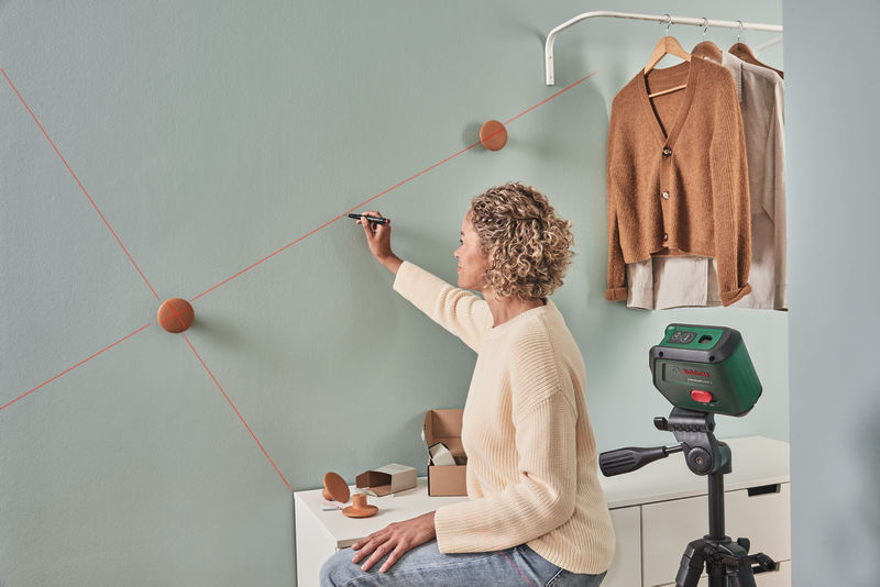 Eine Frau nutzt einen Laserpointer auf einem Stativ, um Linien an eine grüne Wand zu zeichnen.