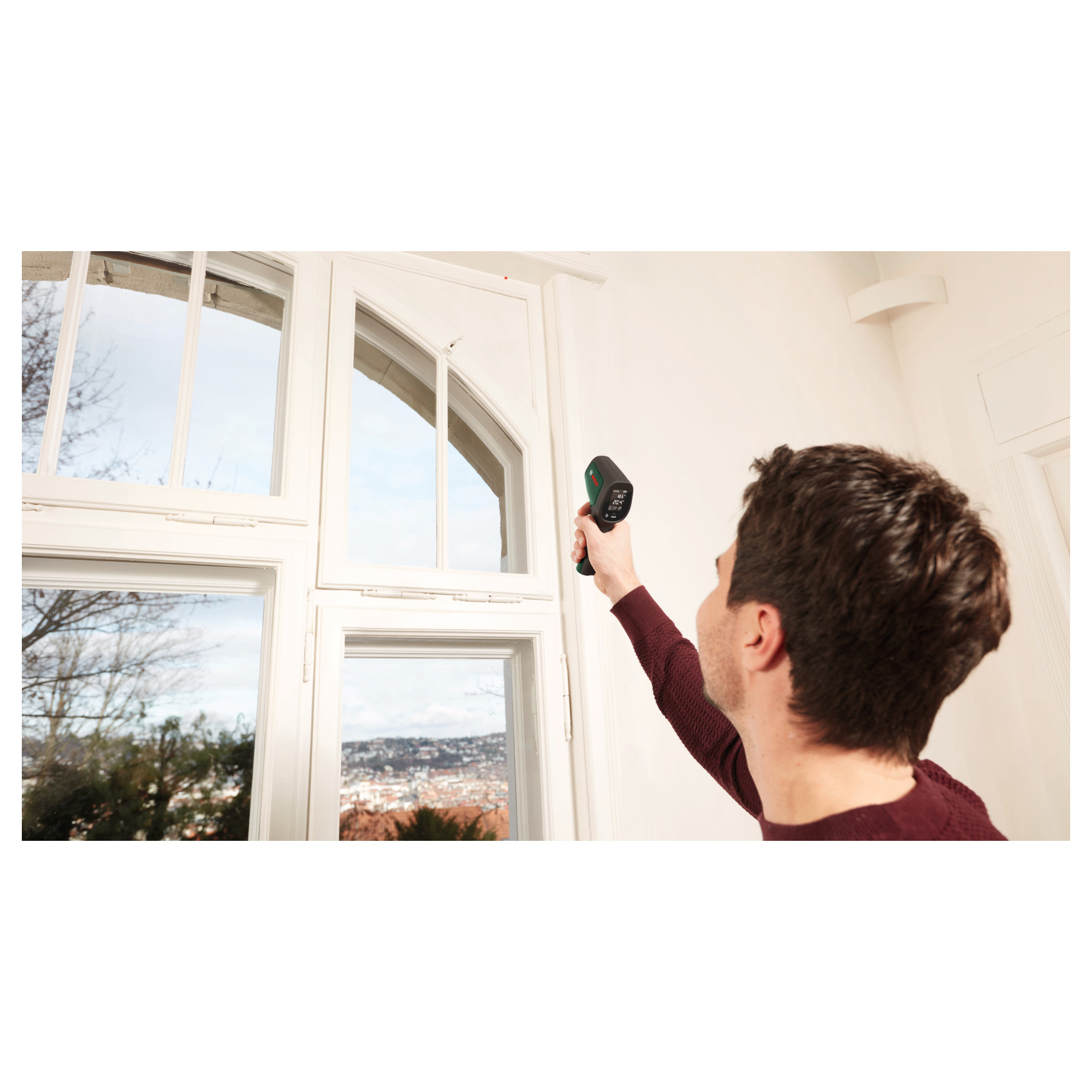 Ein Mann im roten Pullover misst die Temperatur am weißen Fensterrahmen mit einem Infrarotthermometer.