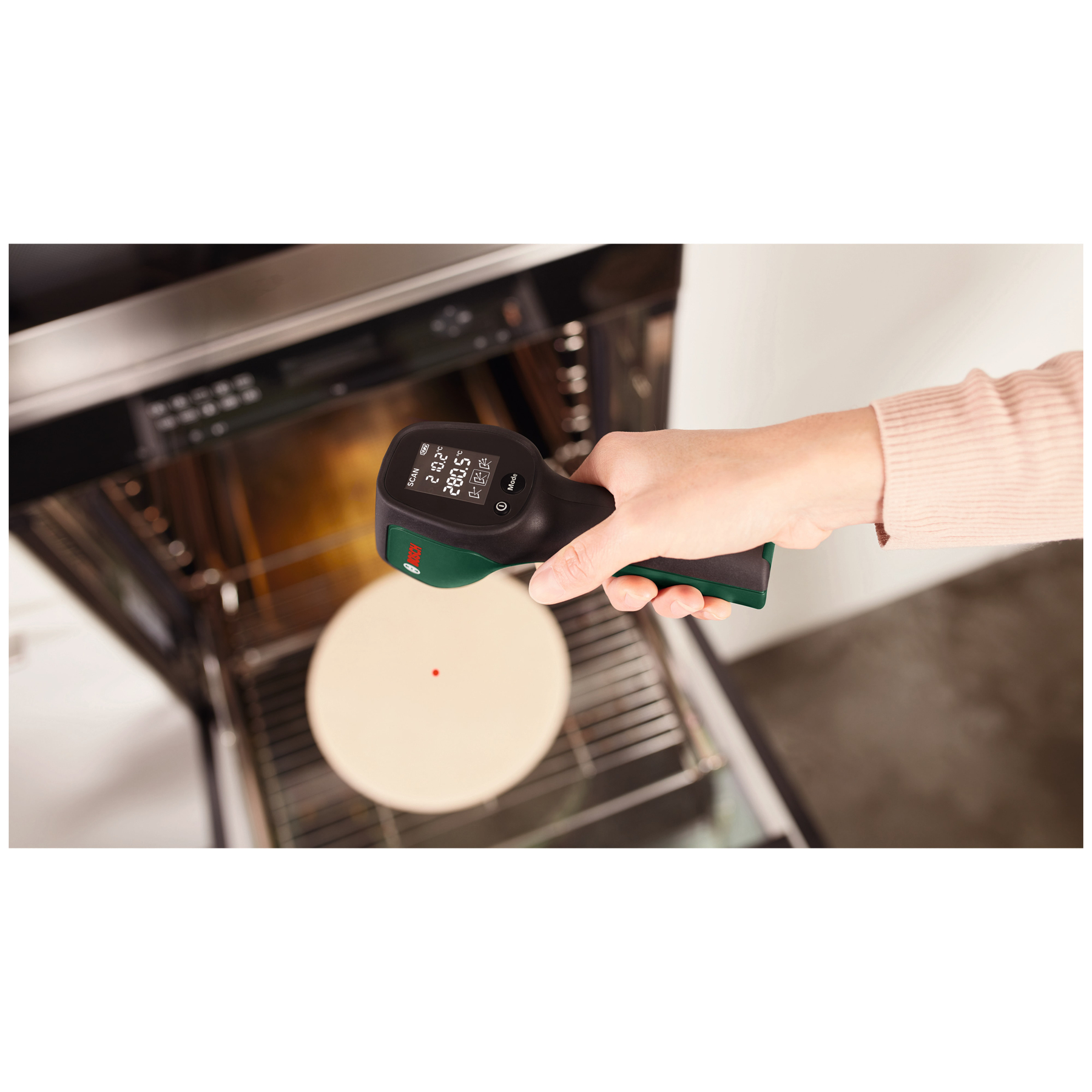 Hand hält Infrarotthermometer vor einen Backofen mit Pizzastein.