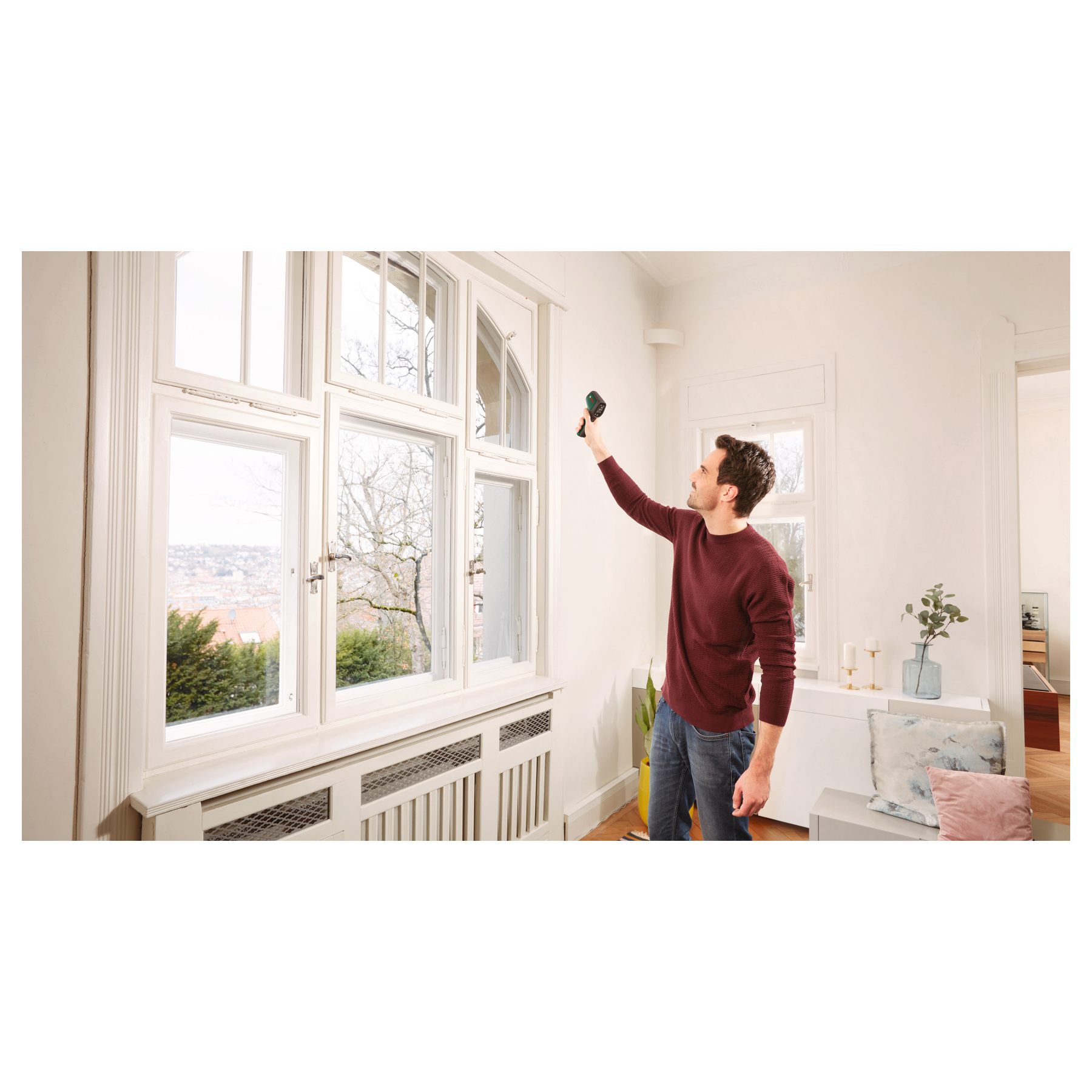 Ein Mann im roten Pullover misst mit einem Messgerät die Temperatur an der Wand neben einem großen weißen Fenster.