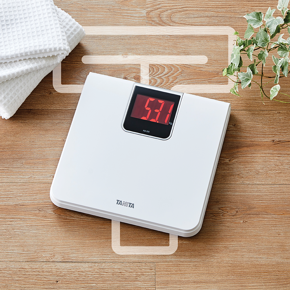A white Tanita scale with a red digital display on a wood-grain floor, next to folded white towels and a green plant.