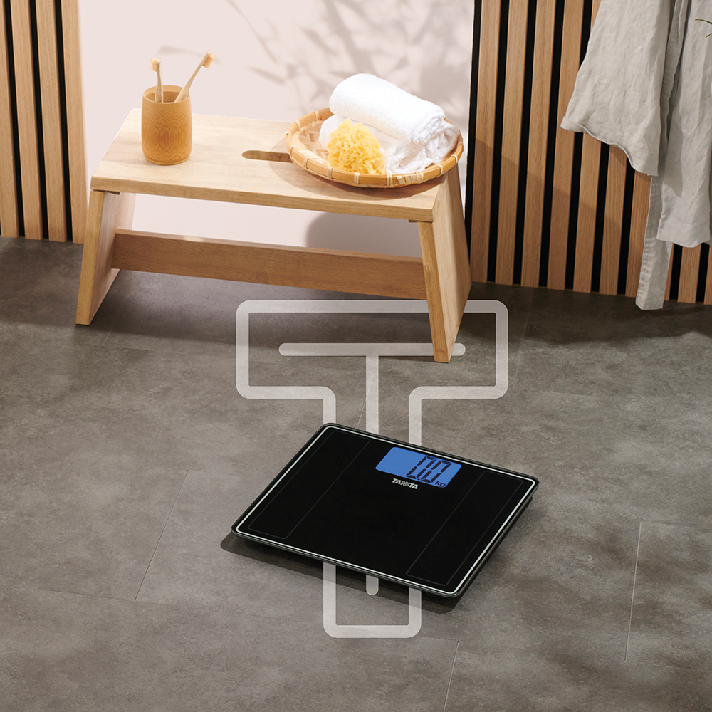 A black digital bathroom scale with a blue display on grey floor, with a wooden stool and towels in the background.