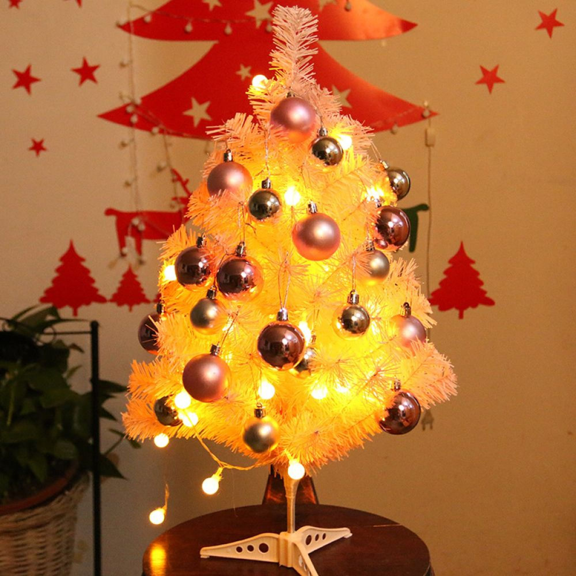 Ein kleiner weißer Weihnachtsbaum mit rosa und silbernen Kugeln und warmen Lichtern steht auf einem Holztisch.