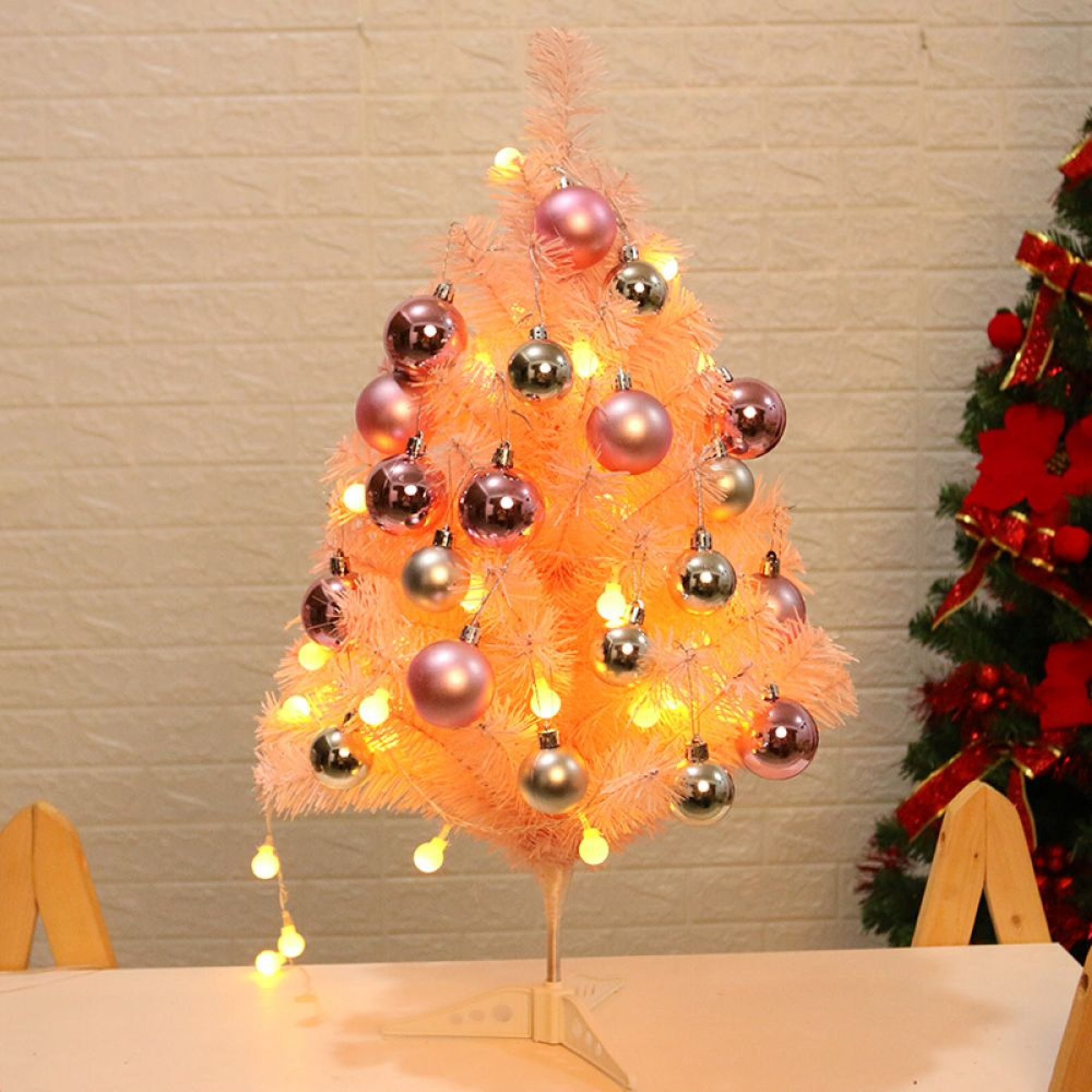 Ein kleiner rosa Weihnachtsbaum mit rosa und silbernen Kugeln und warmen Lichterketten auf einem Tisch.